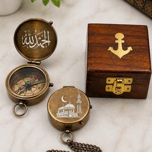 May include: Vintage brass compasses with detailed engravings, featuring Arabic script and a mosque design. A wooden box with a gold anchor emblem is also shown. The compasses have a chain and ring attachment.