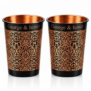 Op de afbeelding: Twee cilindrische metalen bekers met een zwarte buitenkant, een koperkleurig ingewikkeld damastpatroon en de tekst George & honey bij de rand. De binnenkant van elke beker heeft een gepolijste koperen afwerking met een gladde textuur.