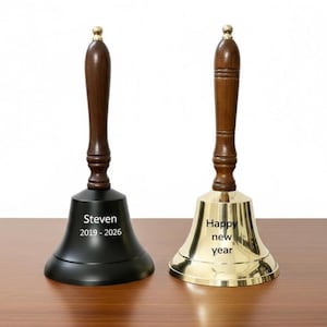 Puede incluir: Dos campanas de mano con mangos de madera y detalles de latón. Una campana es negra con el nombre "Steven" y los años "2019-2026" grabados. La otra campana es dorada con las palabras "Happy new year" grabadas.