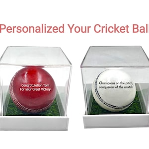 Pode incluir: Duas bolas de críquete em vitrines transparentes. Uma é vermelha com costuras brancas e o texto "Congratulation Tom For your Great Victory." A outra é branca com costuras pretas e o texto "Champions on the pitch, conquerors of the match."