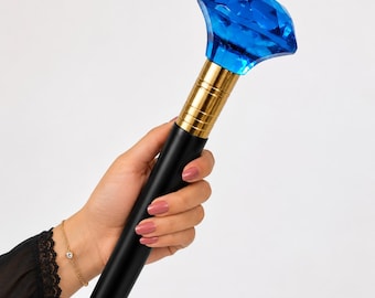 May include: A walking stick featuring a large, faceted, translucent blue diamond-shaped topper made of acrylic. The handle section is connected to a smooth, matte black shaft by a polished gold-colored metal collar with horizontal ribbed detailing.