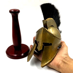 May include: A miniature gold-coloured metal Spartan helmet featuring a prominent black crest made of stiff bristles. Beside the helmet stands a dark reddish-brown wooden display stand with a cylindrical post and a circular base.