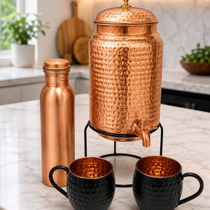 Puede incluir: Un dispensador de agua de cobre martillado con tapa y grifo sobre un soporte de metal negro, una botella de agua de cobre lisa y dos tazas martilladas negras con interior de cobre. El conjunto presenta acabados metálicos y diseño cilíndrico.