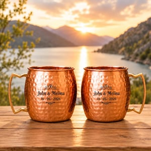 May include: Two hammered copper mugs with gold-toned angular handles. Each mug features black engraved text reading John & Melina and the date 16-05-2026, centered between decorative flourishes. The mugs have a metallic finish and a barrel shape.
