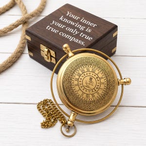 Puede incluir: Una brújula de latón de estilo náutico con una esfera circular que presenta números romanos y anillos concéntricos de números, unida a una cadena dorada. Descansa frente a una caja de madera oscura grabada con el texto: Your inner knowing is your only true compass.