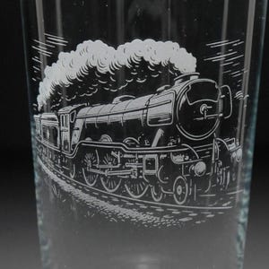 May include: A clear glass featuring an etched image of a steam train, with a cloud of smoke. The design is monochromatic, showing a detailed illustration of the train. This glass could be used for drinking and has a vintage aesthetic.