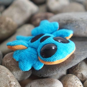 Può includere: Una creatura di peluche blu con grandi occhi neri e macchie nere. Il giocattolo ha dettagli arancioni e poggia su un letto di pietre grigie. Il design della creatura ricorda un personaggio dei cartoni animati.