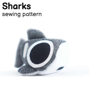 Pode incluir: Um brinquedo de pelúcia de tubarão cinza e branco com um olho preto e detalhes brancos. O tubarão tem uma barbatana nas costas e o texto "Sharks sewing pattern" em preto acima. O brinquedo é um padrão de costura.