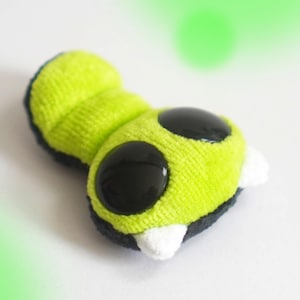Peut inclure: Une peluche en forme de chenille segmentée avec un corps vert citron vif, deux grands yeux circulaires noirs et deux petites protubérances blanches pointues à l'avant. Le dessous du jouet est noir foncé et la texture est douce.