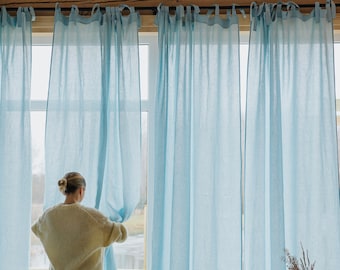May include: A woman stands by a window with light blue sheer curtains. She is wearing a cream sweater and a brown skirt. The window is open and the curtains are blowing in the breeze.