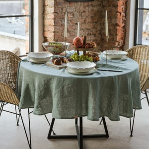 May include: A round table dressed with a pale green linen tablecloth, set for a meal. White bowls and plates are arranged, alongside a wooden serving tray with fruit and pastries. Two wicker chairs are positioned around the table, with a brick wall in the background.