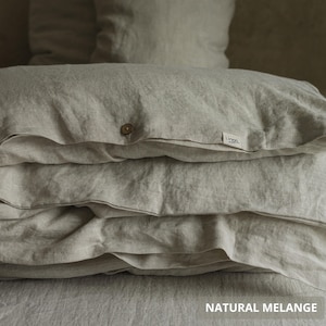 May include: A stack of natural-colored linen bedding, including pillows and a duvet cover. The duvet cover has a wooden button closure and a small tag that reads "Linen." The text "NATURAL MELANGE" is at the bottom of the image. The bedding is folded neatly.