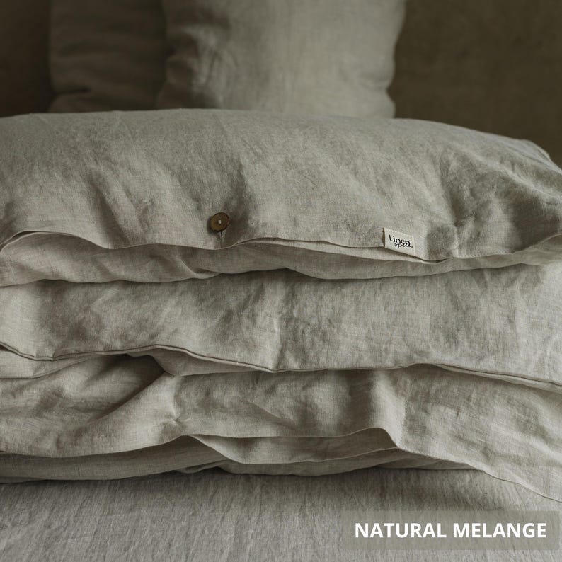 May include: A stack of natural-colored linen bedding, including pillows and a duvet cover. The duvet cover has a wooden button closure and a small tag that reads "Linen." The text "NATURAL MELANGE" is at the bottom of the image. The bedding is folded neatly.