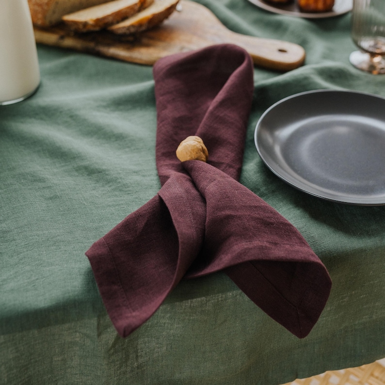 May include: A burgundy linen napkin, folded and held by a wooden napkin ring, rests on a green tablecloth. A dark gray plate is visible in the background, along with a wooden breadboard and a glass.