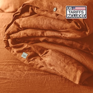 May include: Folded rust-coloured linen duvet cover and sheets. The duvet cover has a button closure and a small white tag with the word "Linen" printed on it. The text "US Tariffs Included" is visible in the upper right corner.