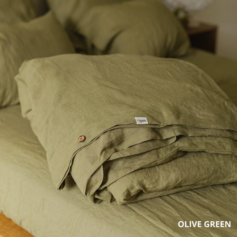 May include: Olive green linen bedding set, including a folded duvet cover with a wooden button closure and a small label. The set is displayed on a bed with matching pillowcases, creating a cohesive and inviting aesthetic.