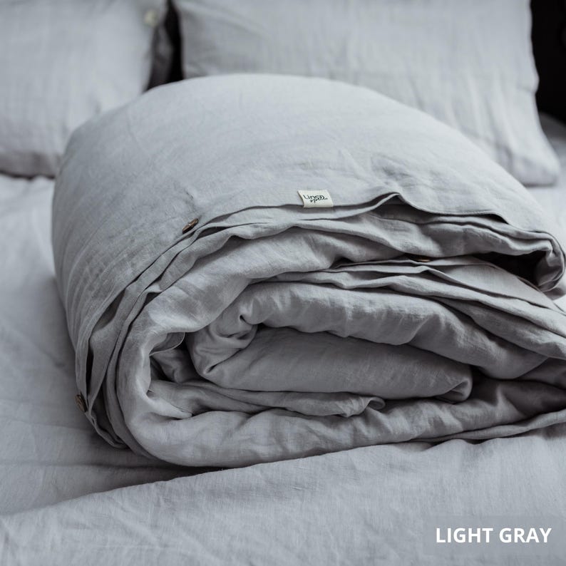 May include: A light gray linen duvet cover folded on a bed, with two matching pillows in the background. The duvet cover has a tag that reads "Linen." The text "LIGHT GRAY" is visible in the bottom right corner.