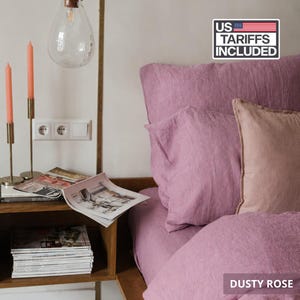 Könnte beinhalten: Ein Bettwäsche-Set aus altrosa Leinen mit einem Bettbezug und zwei Kissen, eines in Altrosa und eines in einem helleren Blush-Ton. Auf einem Nachttisch aus Holz liegen Zeitschriften, zwei hohe orangefarbene Stabkerzen und eine Glas-Hängeleuchte.