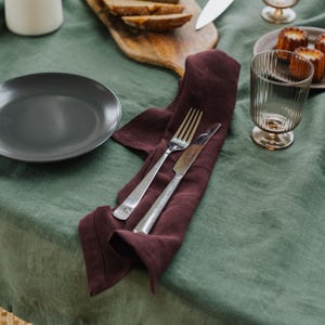 May include: A dining table setting with a dark gray plate, a burgundy napkin with a fork and knife, and a clear glass. The table is covered with a green tablecloth, and there is a wooden cutting board with bread.