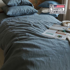 Peut inclure: Un lit est fait avec une literie en lin bleu-gris, comprenant une housse de couette et plusieurs oreillers. Un magazine est ouvert sur le lit. La scène est bien éclairée, suggérant un cadre de chambre confortable et accueillant. Le logo US Tariffs Included est visible.