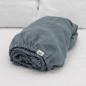 Könnte beinhalten: Ein gerolltes, staubblaues Spannbettlaken. Das Leinentuch hat einen elastischen Rand. Ein kleines, rechteckiges Etikett mit der Aufschrift "Linen Spells" ist angebracht. Das Laken liegt auf einer weißen Oberfläche.