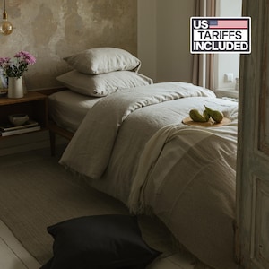 May include: A bed dressed in light beige linen bedding, including two stacked pillows and a duvet cover. A cream-colored throw blanket is draped over the foot of the bed, topped with two green pears on a small wooden plate. A black pillow rests on the floor.