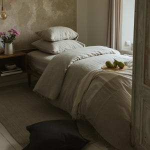 May include: A bed with light beige bedding, including a duvet, pillows, and a blanket. Two pears rest on a wooden tray on the bed. A black pillow is on the floor. A small wooden nightstand holds a vase of flowers.