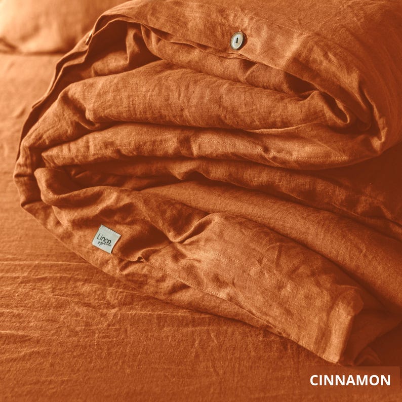 May include: A folded, rust-colored linen duvet cover with a button closure. The bedding is neatly stacked, showcasing the texture of the fabric. The word "Cinnamon" is displayed at the bottom right corner of the image.