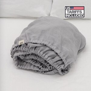 Peut inclure: Un drap-housse gris clair plié avec des bords élastiques, posé sur une surface blanche. Le drap porte une étiquette "Linen Spells". L'image comprend un graphique "US Tariffs Included".