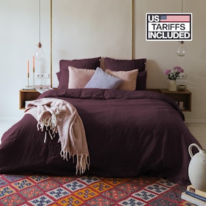 Könnte beinhalten: Ein tiefvioletter Leinen-Bettbezug auf einem Bett mit passenden Kissenbezügen, zwei hellrosa Kissen und einem hellblauen Kissen. Eine strukturierte hellrosa Decke mit Fransen liegt am Fußende des Bettes. Der Text im Bild lautet US TARIFFS INCLUDED.