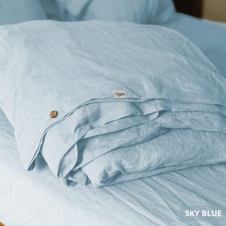 May include: Sky blue linen bedding set, folded neatly on a bed. The set includes a duvet cover and possibly pillowcases, with a wooden button detail. The fabric has a soft, textured appearance, suggesting comfort and quality.