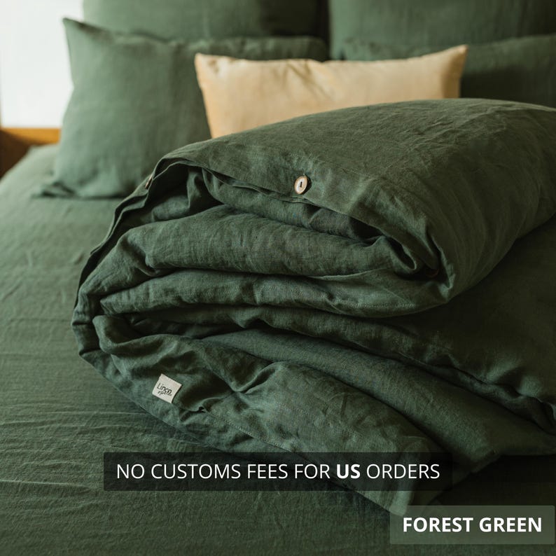 May include: Forest green linen bedding set, including a duvet cover, pillowcases, and fitted sheet. The duvet cover is folded, showing a wooden button closure. The text "NO CUSTOMS FEES FOR US ORDERS" and "FOREST GREEN" are visible.