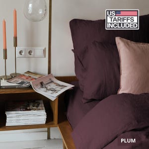 May include: A bedding set featuring a deep plum-colored duvet cover and pillowcases paired with a single light pink accent pillow. The bed is positioned next to a wooden nightstand holding a stack of magazines and two tall peach-colored candles.