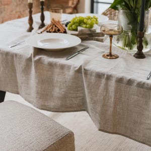 Könnte beinhalten: Eine beige Leinentischdecke bedeckt einen Tisch, der für eine Mahlzeit gedeckt ist. Ein weißer Teller, Besteck, ein Glas und ein Stielglas stehen auf dem Tisch. Grüne Trauben und Brotstangen sind ebenfalls vorhanden. Zwei braune Kerzenständer und eine Vase mit Blumen sind sichtbar.