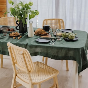 May include: A dining table set for a meal, featuring a dark green linen tablecloth. The table is set with plates, glasses, silverware, and food items like bread, artichokes, and nuts. Wicker chairs surround the table, creating a rustic aesthetic.