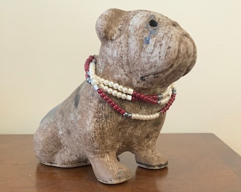 Pode incluir: Uma estatueta de bulldog castanha com superfície texturizada e aspeto envelhecido, usando um colar em camadas feito de pequenas pérolas brancas, contas vermelho-escuras e espaçadores quadrados prateados à volta do pescoço.