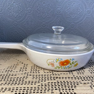 Puede incluir: Plato para hornear blanco con un dise&ntilde;o floral con flores naranjas, azules y amarillas. El plato tiene una tapa de vidrio transparente.
