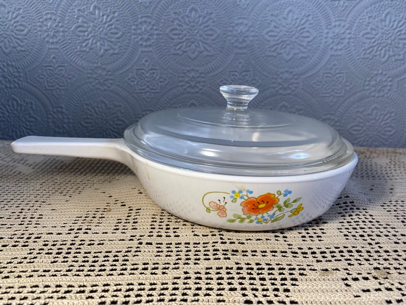 Puede incluir: Plato para hornear blanco con un dise&ntilde;o floral con flores naranjas, azules y amarillas. El plato tiene una tapa de vidrio transparente.