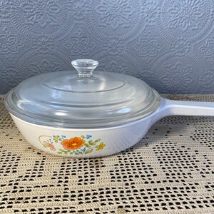 Puede incluir: Plato para hornear blanco con un dise&ntilde;o floral y una tapa de vidrio. El plato tiene un asa blanca.