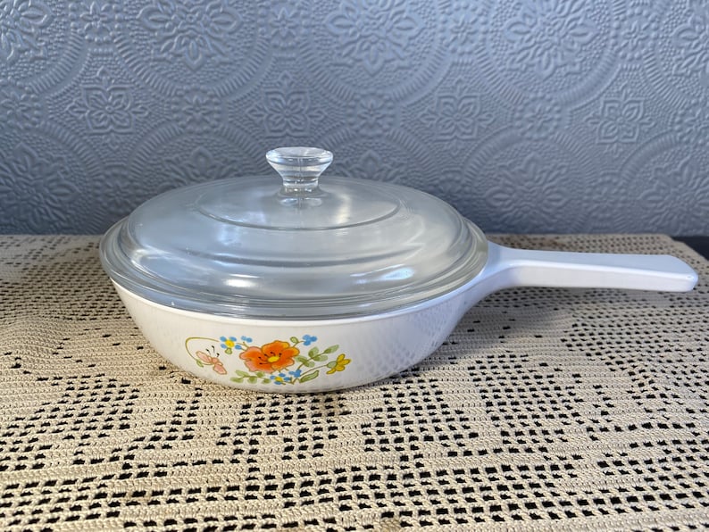 Puede incluir: Plato para hornear blanco con un dise&ntilde;o floral y una tapa de vidrio. El plato tiene un asa blanca.