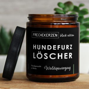 Könnte beinhalten: Ein braunes Glas mit schwarzem Deckel und einem schwarzen Etikett mit weißem Text, der "FRECHEKERZEN black edition" und "HUNDEFURZ LÖSCHER" lautet. Das Glas ist handgefertigt in Berlin und duftet nach einem Waldspaziergang.