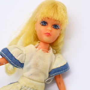 Puede incluir: Una muñeca vintage con cabello rubio y ojos azules. La muñeca lleva un vestido blanco con ribetes azules en las mangas. El brazo de la muñeca es naranja con algunas manchas oscuras. Un juguete clásico.