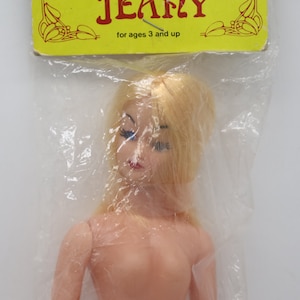May include: A plastic doll with blonde hair and blue eyes. The doll is packaged in a clear plastic bag with a yellow label that reads "Jeanie for ages 3 and up".