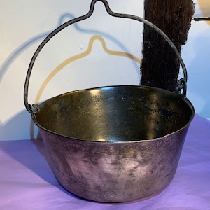 May include: A large, round, copper pot with a metal handle. The pot has a worn, aged look and is sitting on a purple surface.