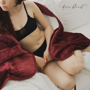 May include: A woman is sitting on a white couch wrapped in a burgundy fleece blanket. The blanket has a sherpa lining. The woman is wearing a black bra and black underwear.