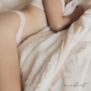 May include: A person lying in bed with a white blanket. The blanket is soft and fluffy. The person is wearing white underwear. The text "free bleed" is visible in the lower right corner of the image.