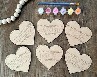 Puede incluir: Seis adornos de madera en forma de corazón sin terminar con diferentes frases pintadas en ellos: "Be Mine", "Hug Me", "XOXO", "I Luv U", "Riley" y "Cutie Pie".