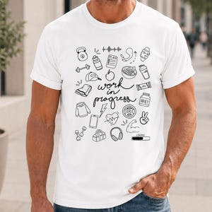 Könnte beinhalten: Ein weißes kurzärmeliges T-Shirt mit einem schwarzen Grafikdruck verschiedener Fitness-Symbole, darunter eine Kettlebell, Hanteln, eine Wasserflasche, ein Herz und Kopfhörer, mit dem Text "work in progress" in der Mitte.