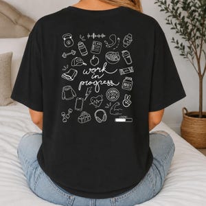 Könnte beinhalten: Ein schwarzes T-Shirt mit einem weißen Grafikdruck auf der Rückseite. Das Design zeigt den Text "work in progress", umgeben von verschiedenen Fitness-Symbolen wie einer Kettlebell, Hanteln, einer Trinkflasche, einem Herz, Kopfhörern und einem Ladebalken.