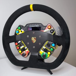 May include: A black suede racing steering wheel with a yellow top center marker. The center console features a Porsche logo, a Fanatec display, and multiple colored buttons labeled with functions like Radio, Starter, Ign, T/C, Map, Abs, and Pit Speed.